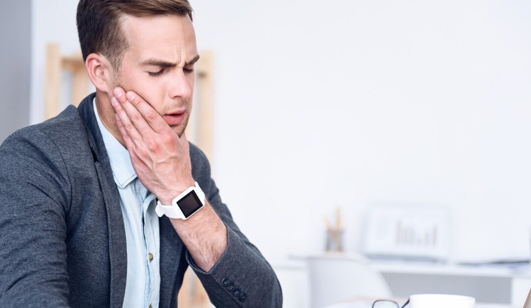 A man wearing a sports jacket sits at a desk with a laptop, glasses, and a white mug. He holds his jaw and winces in pain.