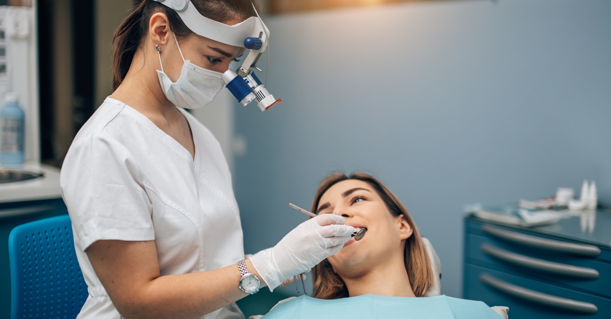 In an exam room, a female dentist is wearing a dental loupe and looking at the patient's teeth using a small mouth mirror.