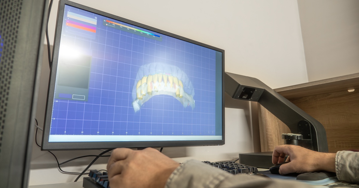 A person sits at a desk. A computer monitor features a 3D model of a patient's teeth using computer-aided design technology.
