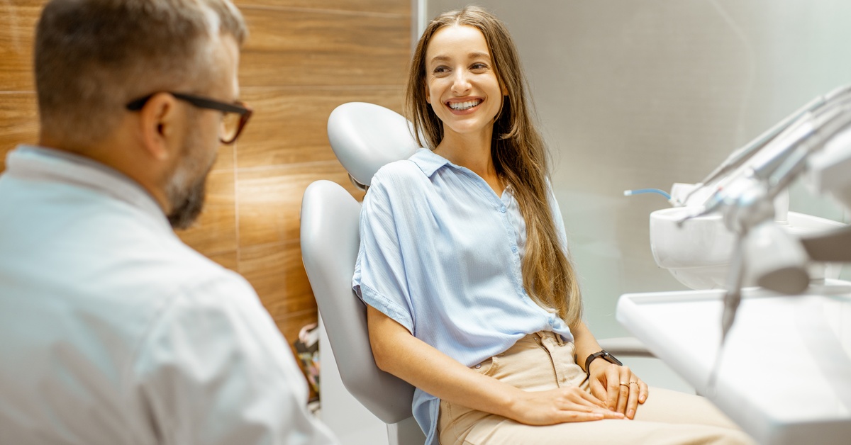 A woman sits in a dental chair in an exam room. She looks and smiles at a dentist wearing a white coat across from her.