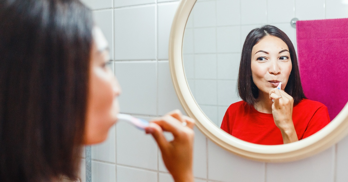 A woman wearing a red shirt looks into a round mirror as she brushes her teeth. The wall is made of white tiles.