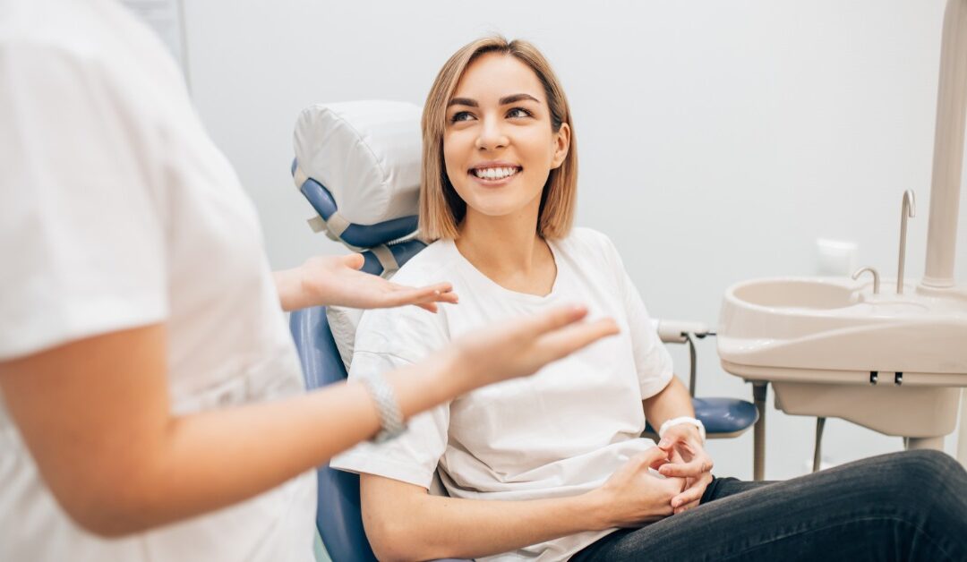 A young woman sits upright with her hands folded in a dental exam chair. The dentist is standing and speaking to her.