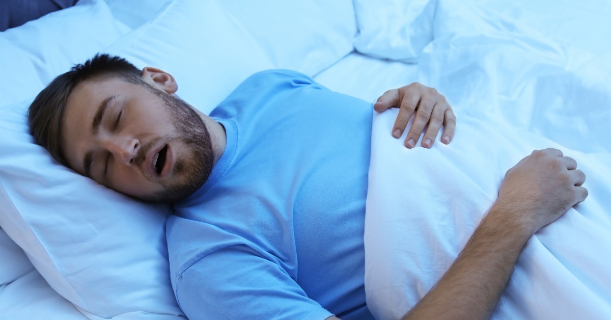 A man wearing a blue shirt is lying on his back in a bed. He's sleeping with his mouth open and clutching a white comforter.