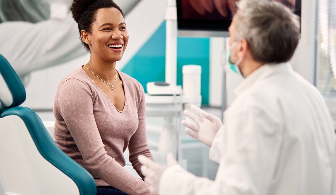 A woman is sitting sideways in a dental exam chair and facing a dentist. The dentist is wearing gloves and a mask.