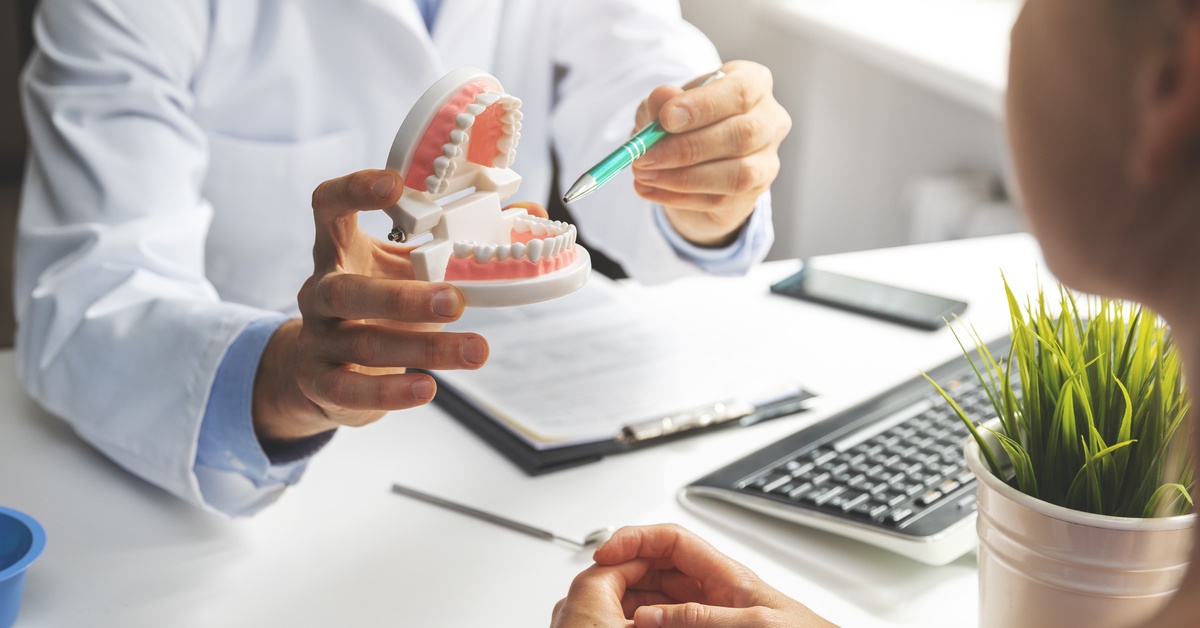 A dentist is sitting across from a patient. The dentist is holding a mold of teeth and gums while pointing to a tooth.