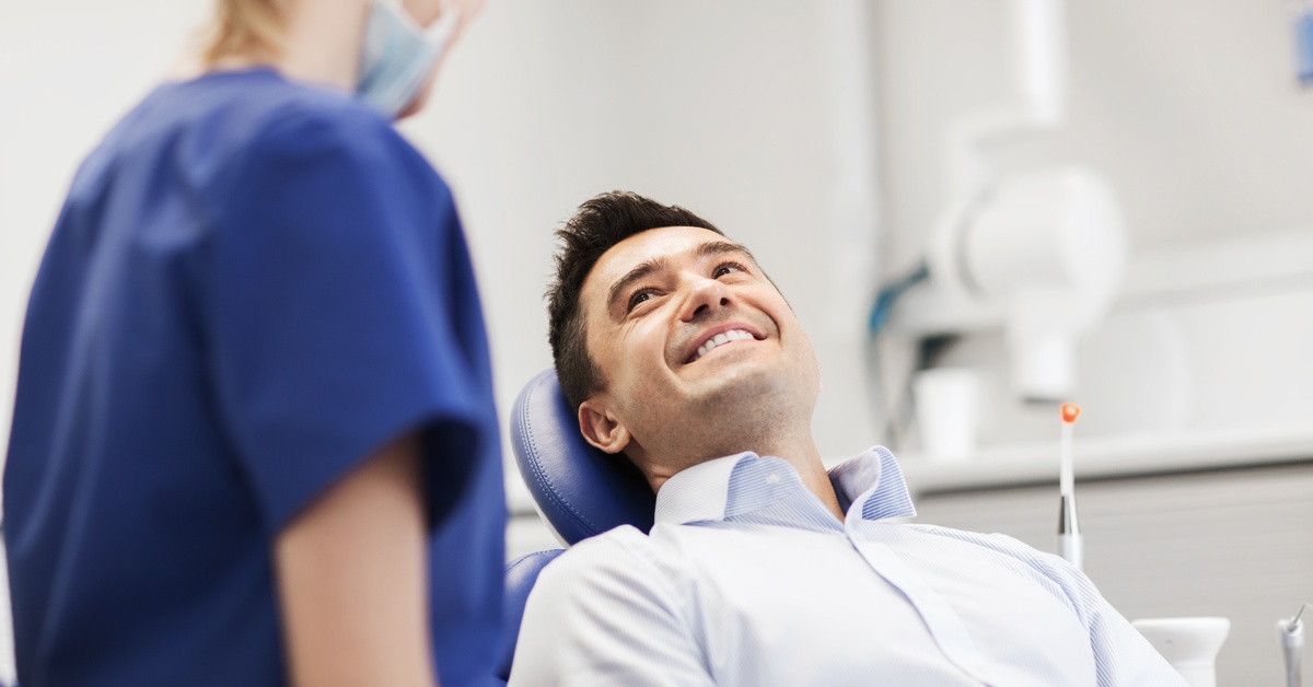A man lies back on a dark blue dental exam chair. He smiles and looks at a dental hygienist wearing blue scrubs.