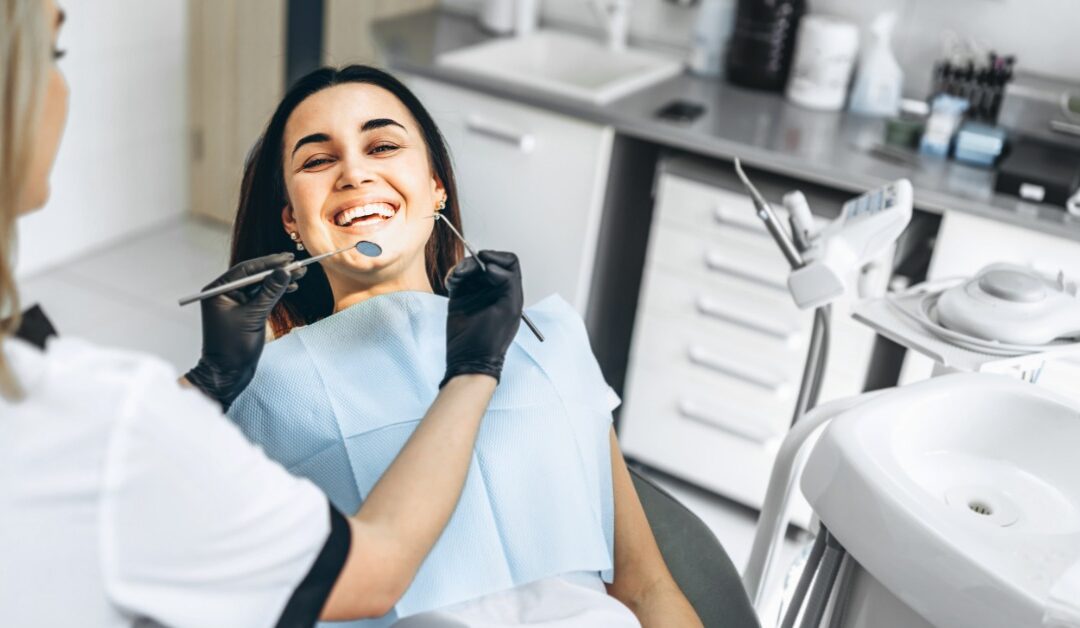 A close-up of a woman sitting in a dental exam chair and smiling. A dentist wears a mask and gloves while holding two tools.