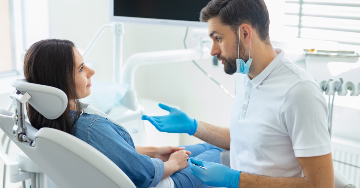 An oral hygienist wearing blue gloves speaks to a female patient sitting back in a chair in an exam room.