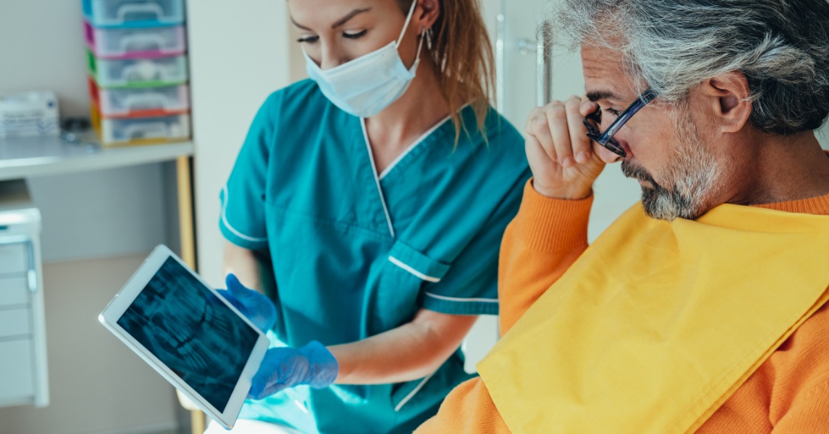 A woman wearing scrubs and a mask holds a tablet showing an X-ray of a patient's teeth. The male patient looks a the screen.