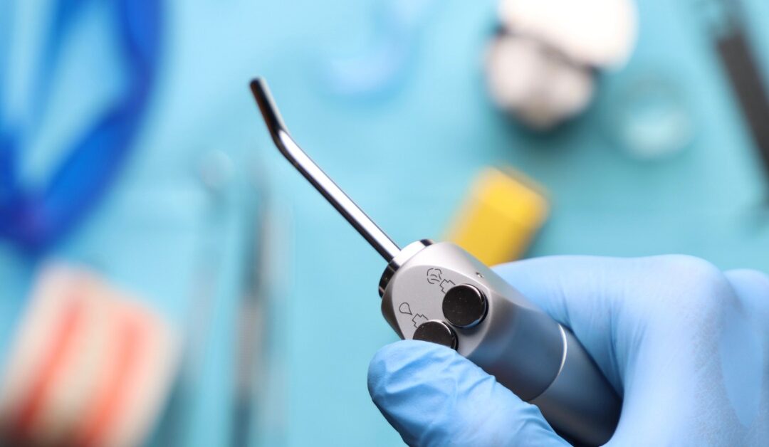 An extreme close-up of a gloved hand holding an oral irrigator above a tray. The blue tray has various dental tools.