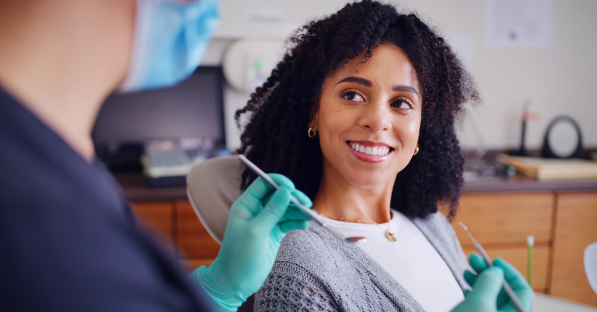 A woman is sitting upright in a dental chair and smiling at a dentist. The dentist wears gloves and a mask.