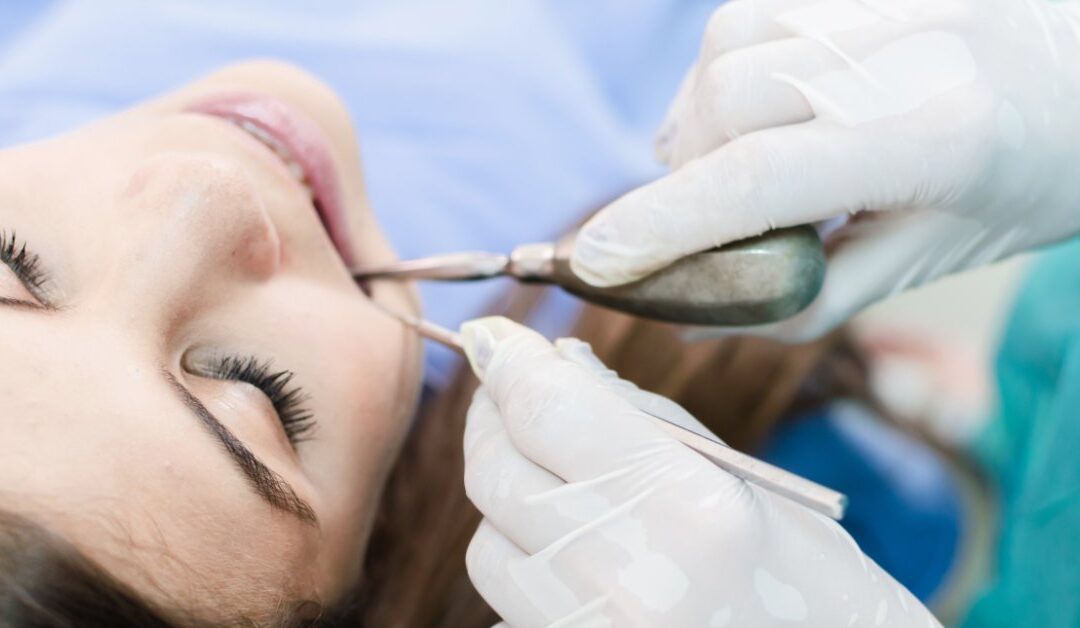 A young woman is lying in a chair while a dentist's gloved hands are using instruments near the patient's open mouth.