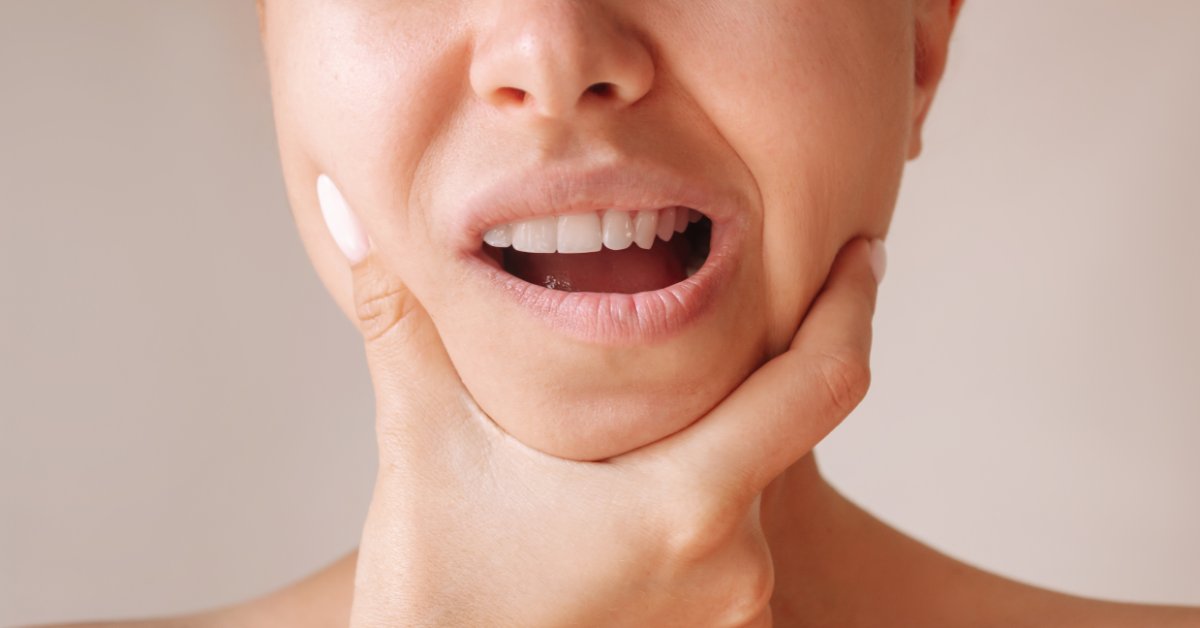 A cropped view of a woman holding her chin with one hand against a plain beige background while looking uncomfortable.