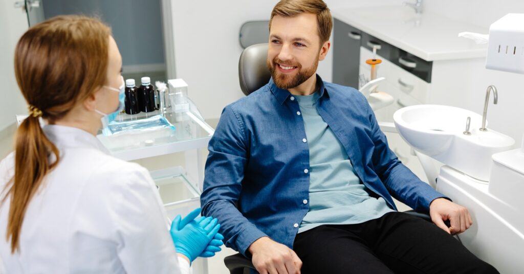 A man sitting in a dental chair while a person in gloves talks with him beside a sink and treatment tools.