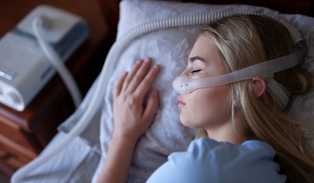 A person lying in bed is wearing a CPAP mask, which is connected to a breathing machine on a nightstand.