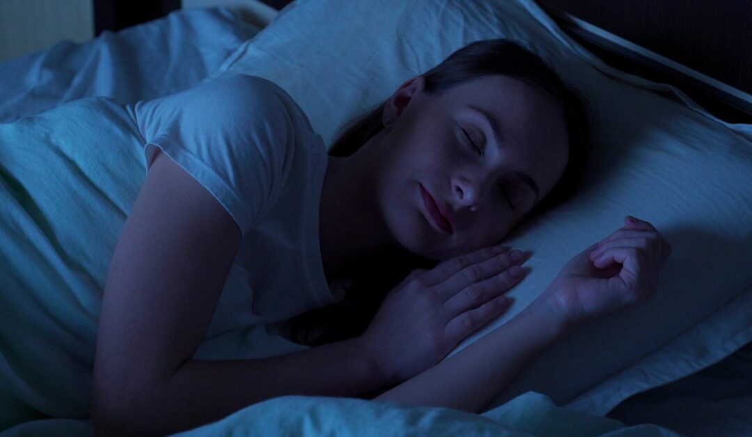 A woman is sleeping on her side and resting her head on a pillow. The sheets are white, and the room has a blue tint.