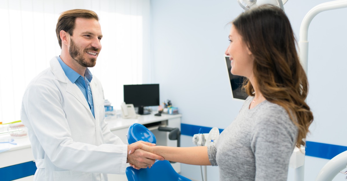 A smiling man in a white coat shaking hands with a woman beside a blue chair in a dental office with overhead equipment.