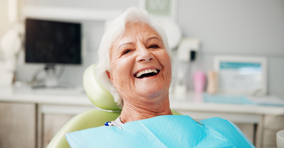 A senior adult with white hair is sitting in a green dental chair. She's wearing a blue bib in a bright dental office.