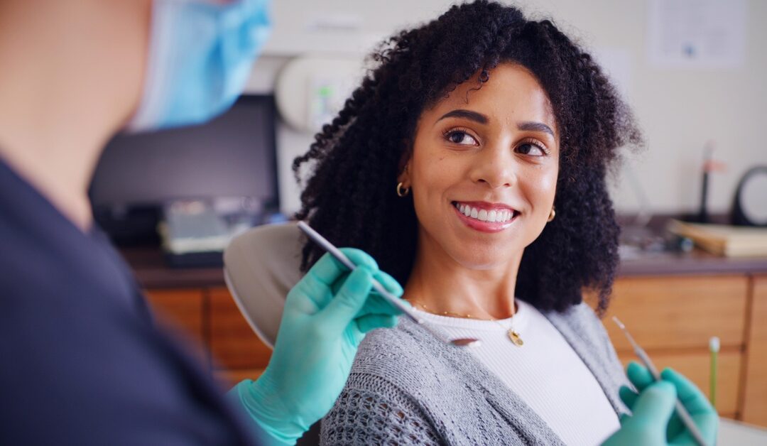 A smiling woman sits in a dental chair while a masked person wearing green gloves holds metal dental tools.