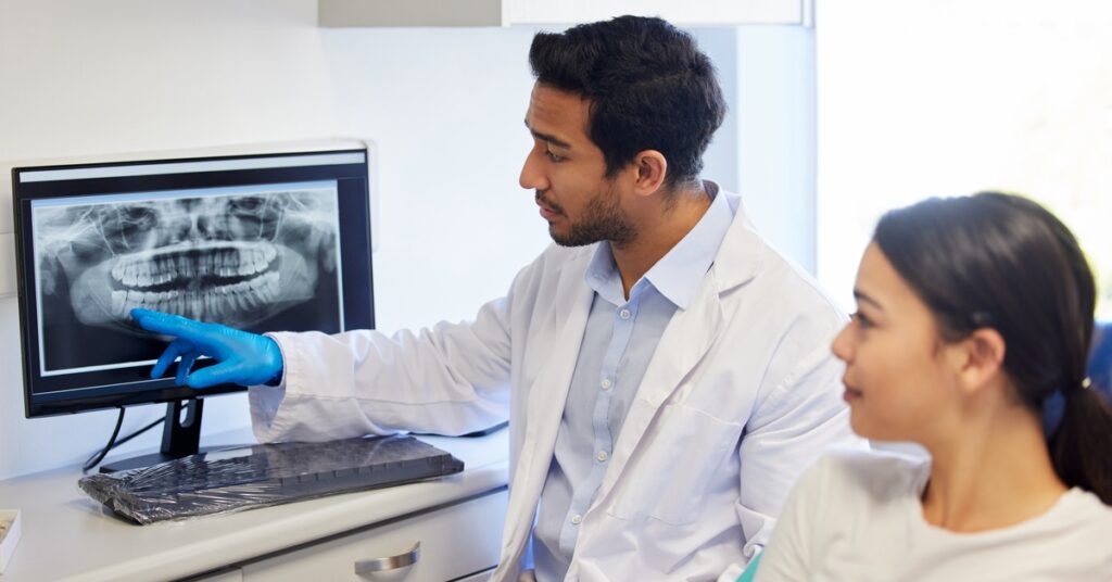 A dentist wearing a white coat points at a dental X-ray on a computer screen. A woman sits beside him in the office.