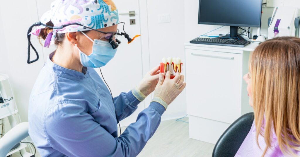 A masked dentist in blue scrubs is holding a dental implant model in front of a seated patient in an exam room.