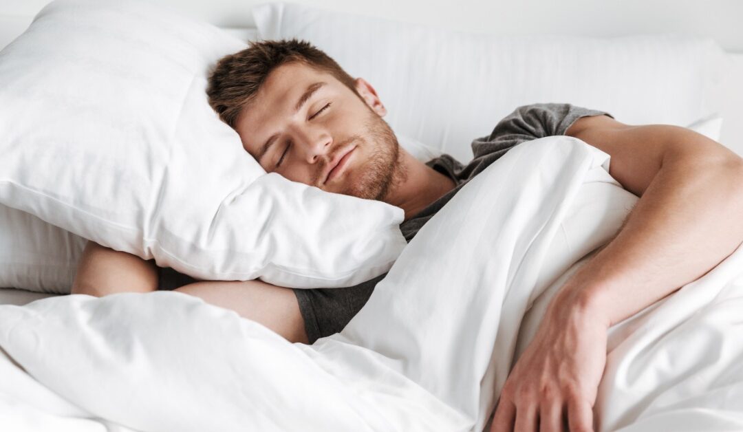 A man sleeping on a white pillow under white blankets, with one arm resting outside the bedding in a bright bedroom.