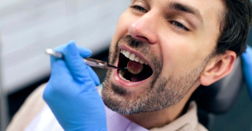 A person wearing blue gloves holding a dental mirror inside a man's open mouth while he sits in a dental chair.
