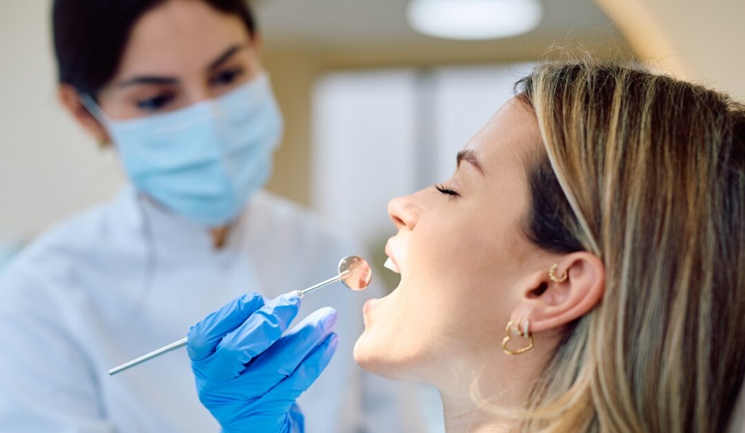 A masked person in blue gloves holding a dental mirror near a woman with an open mouth in a dental chair.