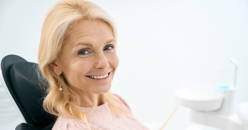 A smiling blonde woman wearing a pink sweater is sitting upright in a black dental chair. Equipment fills the background.