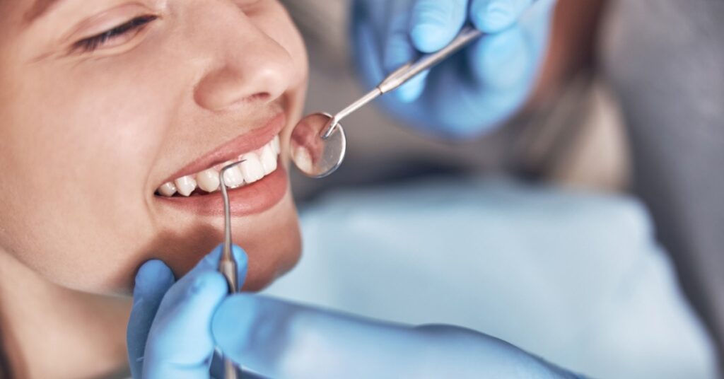 A person wearing blue gloves uses a dental mirror and tool to closely examine the teeth of a smiling woman.