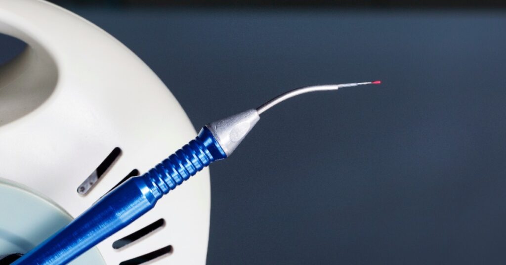 A blue handheld dental laser instrument with a thin curved metal tip and a tiny red light beside a white equipment housing.