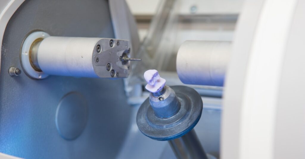 A small white dental crown attached to a tilted metal holder inside a milling machine between two cylindrical cutting tools.